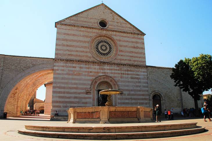 Zicht op de gevel van de Sint Chiara kerk in Assisi Umbrië Italië. Het roosvenster en de karakteristieke bogen aan de zijkant van de kerk zijn zichtbaar. Op de voorgrond de achthoekige fontein.