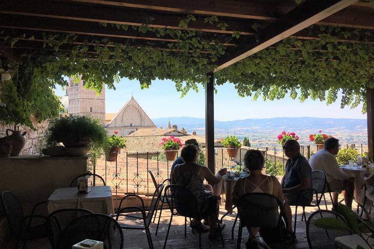 Mensen genieten van een lunch in de schaduw met uitzicht op het lager gelegen Assisi. Op de foto is ook een stuk van de Sint Rufino kathedraal te zien.